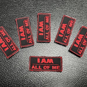 Könnte beinhalten: Sechs schwarze rechteckige Aufnäher mit rotem gesticktem Text "I AM ALL OF ME". Die Aufnäher sind auf einer strukturierten schwarzen Oberfläche angeordnet.