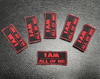 May include: Six black rectangular patches with red embroidered text that reads "I AM ALL OF ME". The patches are arranged on a textured black surface.