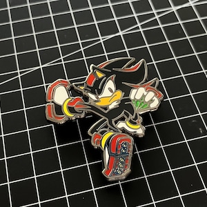 May include: A detailed enamel pin featuring the character Shadow the Hedgehog from the Sonic the Hedgehog video game series. The pin is primarily black with red, yellow, and white accents. The character is in a dynamic pose.