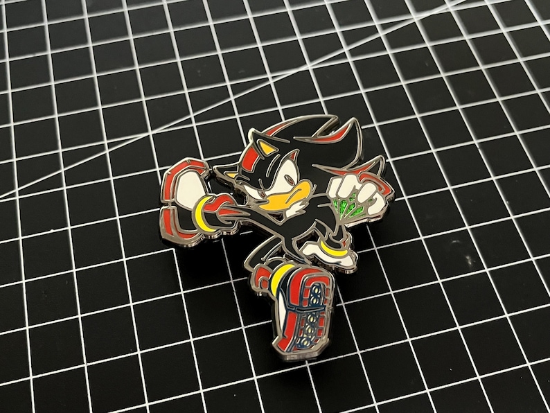 May include: A detailed enamel pin featuring the character Shadow the Hedgehog from the Sonic the Hedgehog video game series. The pin is primarily black with red, yellow, and white accents. The character is in a dynamic pose.
