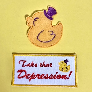 May include: Yellow duck patch with a purple top hat and the text "Take that Depression!" in red script on a white rectangle with a yellow border. The duck is wearing a purple top hat.