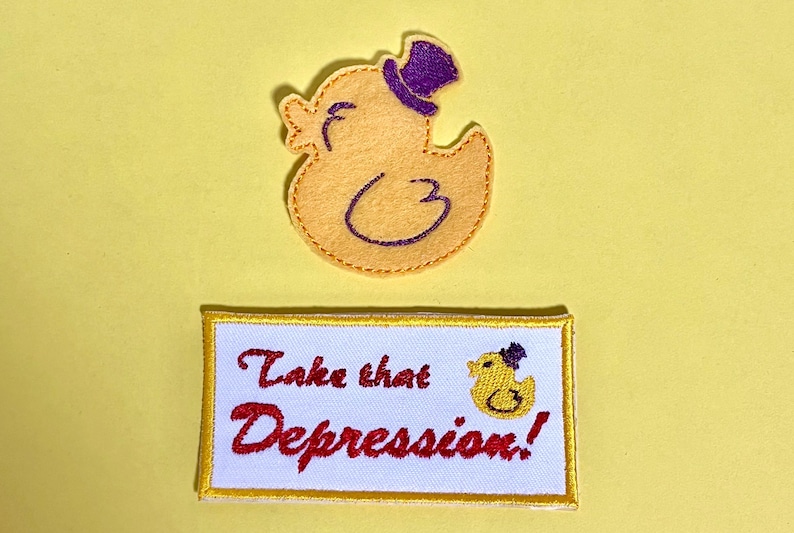 May include: Yellow duck patch with a purple top hat and the text "Take that Depression!" in red script on a white rectangle with a yellow border. The duck is wearing a purple top hat.