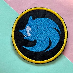 May include: Embroidered patch with a blue character design on a black circular background. The character has a white mouth and is surrounded by a yellow border. Suitable for clothing or accessories.