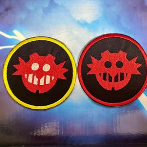 Op de afbeelding: Twee ronde patches met een rood, gestileerd gezicht met een grijnzende mond op een zwarte achtergrond. Eén patch heeft een gele rand, de andere een rode rand. Deze patches zijn waarschijnlijk voor kleding of accessoires.