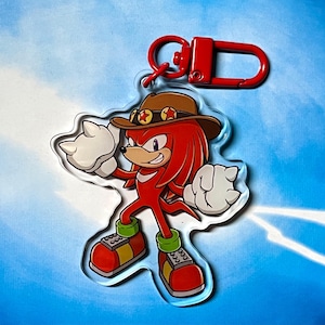 May include: A clear acrylic keychain featuring a cartoon character with red fur, white gloves, and green and red shoes. The character wears a brown cowboy hat with star accents. A red metal clasp is attached to the top.