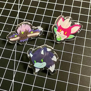 May include: Three acrylic pins featuring cartoon characters. One pin depicts a moth with purple and brown wings. Another shows a pink and white fox-like character. The third pin is a blue werewolf with green eyes.