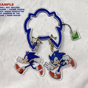 K&ouml;nnte beinhalten: Ein Satz Acryl-Schl&uuml;sselanh&auml;nger mit der Figur Sonic the Hedgehog in zwei verschiedenen Posen. Die Schl&uuml;sselanh&auml;nger sind blau, wei&szlig; und rot, mit einem blauen, wolkenf&ouml;rmigen Aufsatz und einem gr&uuml;nen Edelstein-Anh&auml;nger. Der Text "EXAMPLE" ist sichtbar.