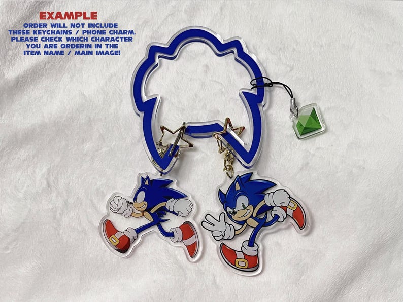 K&ouml;nnte beinhalten: Ein Satz Acryl-Schl&uuml;sselanh&auml;nger mit der Figur Sonic the Hedgehog in zwei verschiedenen Posen. Die Schl&uuml;sselanh&auml;nger sind blau, wei&szlig; und rot, mit einem blauen, wolkenf&ouml;rmigen Aufsatz und einem gr&uuml;nen Edelstein-Anh&auml;nger. Der Text "EXAMPLE" ist sichtbar.