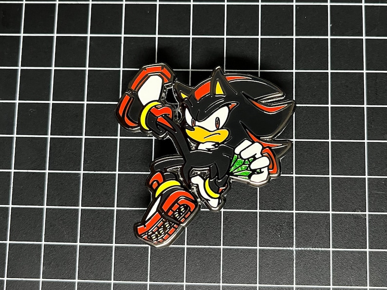 May include: A black, red, yellow, and white enamel pin featuring the character Shadow the Hedgehog from the Sonic the Hedgehog video game series. The pin depicts Shadow in a running pose, holding a green gem. The pin is on a black grid background.