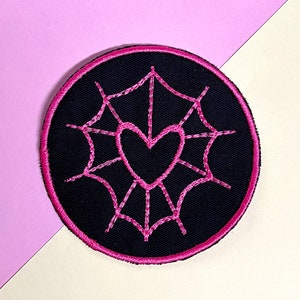 Könnte beinhalten: Ein runder, schwarz gestickter Aufnäher mit einem rosa Spinnennetz-Design und einem Herz in der Mitte. Der Aufnäher hat einen rosa Rand und ist vor einem rosa und weißen Hintergrund platziert.