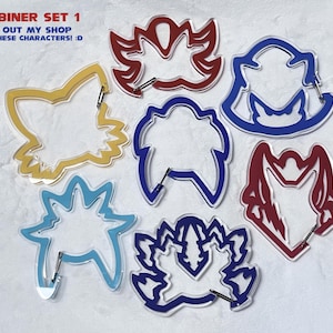 K&ouml;nnte beinhalten: Ein Set aus sieben farbenfrohen Karabinerhaken in verschiedenen Formen, darunter ein Fuchs und florale Designs. Die Karabiner sind gelb, blau und rot umrandet, mit silberner Hardware. Der Text oben lautet "CARABINER SET 1".