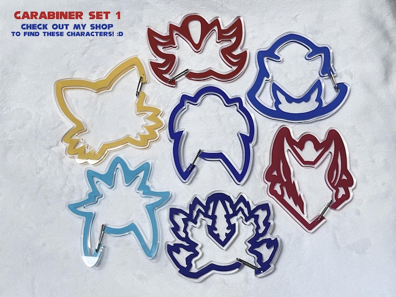 K&ouml;nnte beinhalten: Ein Set aus sieben farbenfrohen Karabinerhaken in verschiedenen Formen, darunter ein Fuchs und florale Designs. Die Karabiner sind gelb, blau und rot umrandet, mit silberner Hardware. Der Text oben lautet "CARABINER SET 1".