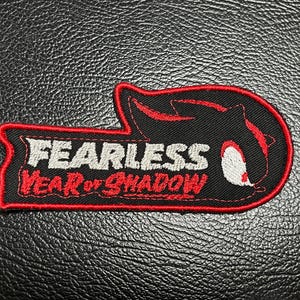 May include: Black and red embroidered patch with the words "FEARLESS Year of Shadow" in white and red. The patch has a unique shape with a red border and a black background.