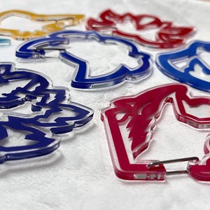 K&ouml;nnte beinhalten: Eine Sammlung farbenfroher Acryl-Schl&uuml;sselanh&auml;nger in Blau, Rot, Gelb und transparent. Jeder Schl&uuml;sselanh&auml;nger hat ein einzigartiges, stilisiertes Design und einen Metallverschluss.