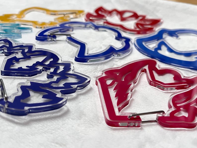 K&ouml;nnte beinhalten: Eine Sammlung farbenfroher Acryl-Schl&uuml;sselanh&auml;nger in Blau, Rot, Gelb und transparent. Jeder Schl&uuml;sselanh&auml;nger hat ein einzigartiges, stilisiertes Design und einen Metallverschluss.