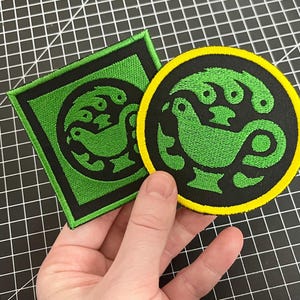 Könnte beinhalten: Zwei gestickte Aufnäher. Einer ist quadratisch mit einem grünen Design auf schwarzem Hintergrund, umrahmt von einem schwarzen Rand. Der andere ist rund, mit einem grünen Design auf schwarzem Hintergrund und einem gelben Rand. Das Design scheint ein stilisierter Vogel zu sein.