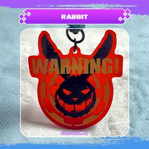 Könnte beinhalten: Ein roter und schwarzer schlüsselanhänger in Hasenform mit dem Wort "WARNING!" in goldfarbenen Buchstaben. Der Schlüsselanhänger hat ein bedrohliches Hasengesicht-Design. Das Wort "RABBIT" steht oben. Der Name des Künstlers, "Sinisters.Art", steht unten.