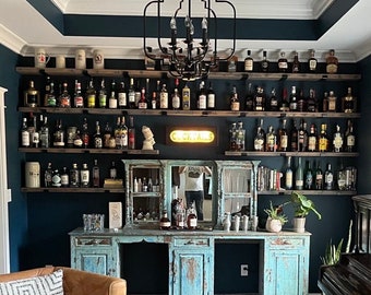 May include: A home bar with a distressed blue cabinet, shelves stocked with liquor bottles, and a black chandelier. The bar is set against a dark blue wall and has a sign that reads "Cocktails".