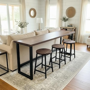 May include: A long, rectangular wooden console table with a dark brown finish and a black metal frame, accompanied by three matching round wooden stools with black metal legs, positioned behind a beige sofa in a bright living room space.