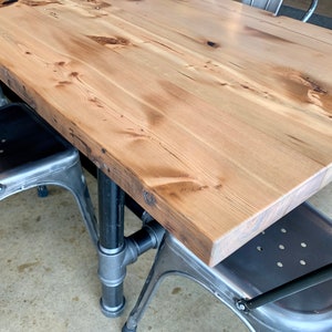 May include: A wooden table top with a natural wood finish and a black metal pipe base. The table is paired with two metal chairs with black metal frames.