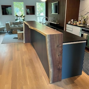 May include: A kitchen island featuring a natural live edge walnut wood countertop that extends down the side panel. The base of the island is finished in a matte dark gray color, contrasting with the warm brown tones of the wood grain.