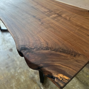 May include: A rectangular table with a live edge slab top made of dark brown walnut wood. The surface displays a prominent natural wood grain pattern, visible knots, and an irregular, organic edge profile. The table is supported by dark metal legs.