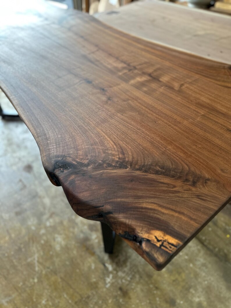 May include: A rectangular table with a live edge slab top made of dark brown walnut wood. The surface displays a prominent natural wood grain pattern, visible knots, and an irregular, organic edge profile. The table is supported by dark metal legs.