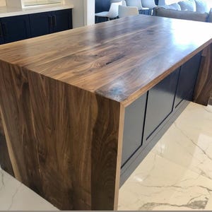 May include: A kitchen island featuring a thick, natural walnut wood countertop with a waterfall edge that extends down the side. The base of the island is constructed with dark navy blue cabinetry panels, providing a contrast to the wood grain.