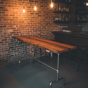 May include: A long, rectangular wooden table with a natural finish, supported by a metal pipe frame. The table is set against a brick wall, with hanging light bulbs and shelves of bottles in the background. The overall aesthetic is rustic and industrial.