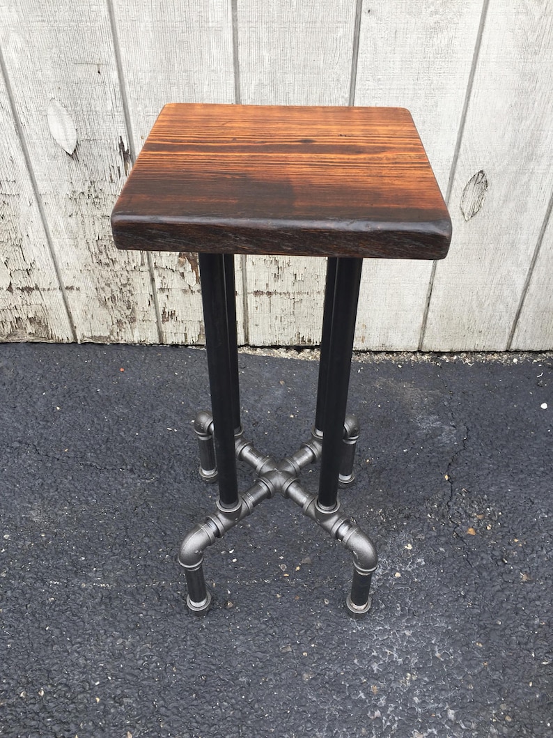 May include: A small, square wooden table with a dark brown wood top and a black metal pipe base. The table is standing on a black tarmac surface.