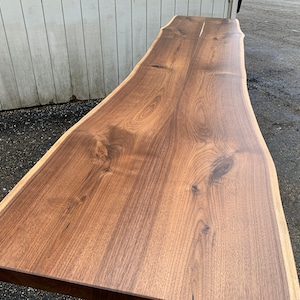 May include: A long, rectangular slab of dark brown walnut wood featuring a natural live edge along the sides. The surface displays a prominent wood grain pattern with visible knots and a smooth, polished finish, showcasing the organic texture.