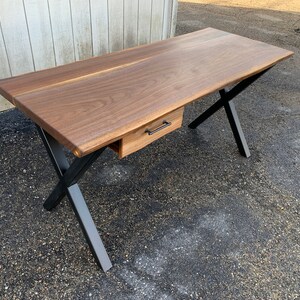 May include: A dark wood desk with a single drawer and black metal X-shaped legs. The desk has a live edge design.