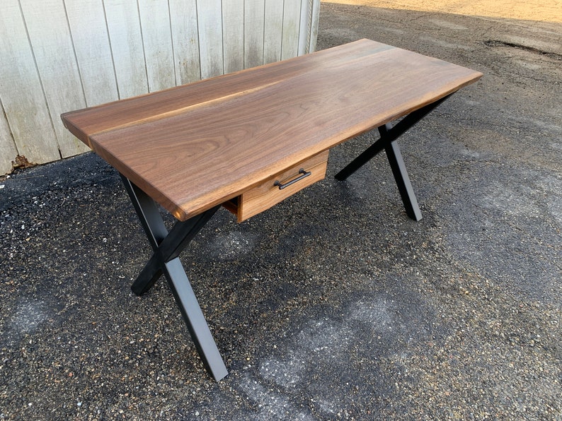 May include: A dark wood desk with a single drawer and black metal X-shaped legs. The desk has a live edge design.