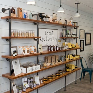 May include: A rustic wood and metal shelving unit filled with various products, including jars of honey, candles, and soaps. The shelves are labeled "Local Apothecary Est. 2005".