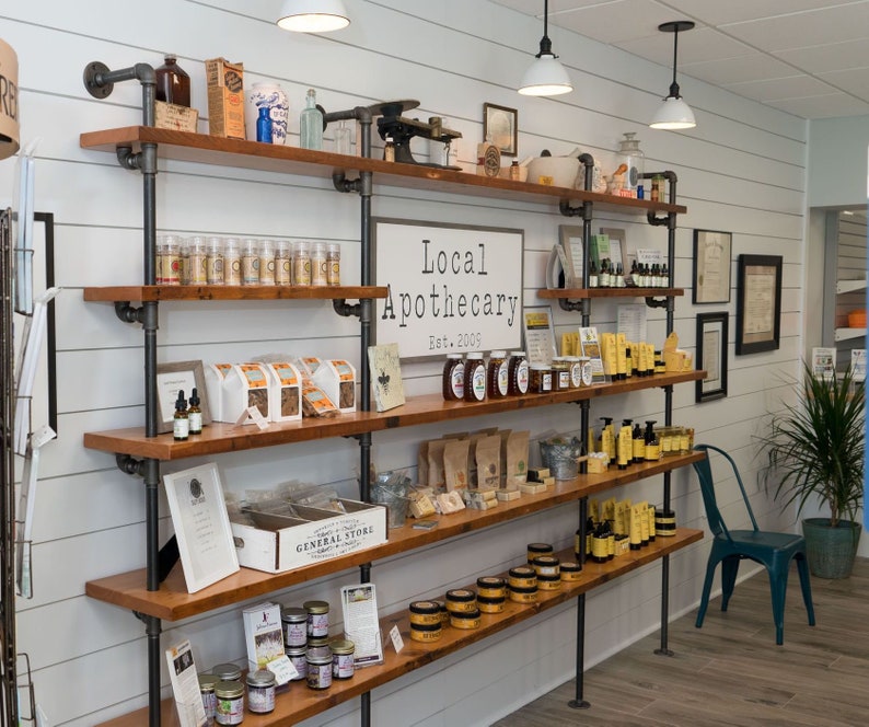 May include: A rustic wood and metal shelving unit filled with various products, including jars of honey, candles, and soaps. The shelves are labeled "Local Apothecary Est. 2005".