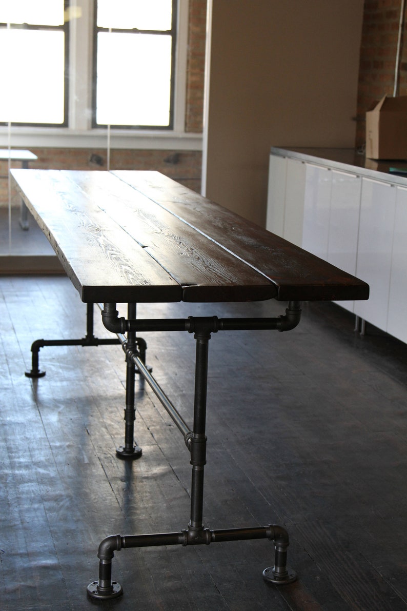 May include: A dark brown, rectangular wooden table with a distressed finish. The table is supported by a black metal pipe frame. The table is approximately 72 inches long and 36 inches wide.