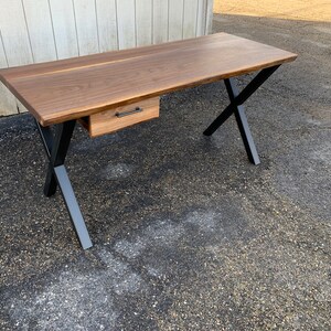 May include: A wooden desk with a single drawer and black metal X-shaped legs. The desk is made of walnut wood and has a natural finish.