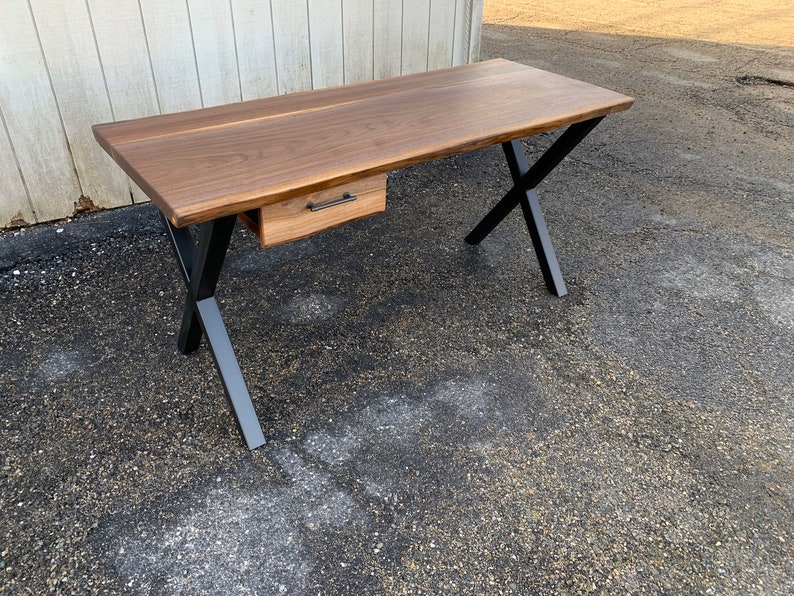 May include: A wooden desk with a single drawer and black metal X-shaped legs. The desk is made of walnut wood and has a natural finish.