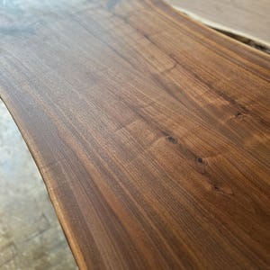 May include: A rectangular wooden slab with a natural live edge, featuring a rich dark brown grain pattern with visible wood knots and a smooth, polished finish. The surface displays deep, warm tones and intricate organic wood textures.
