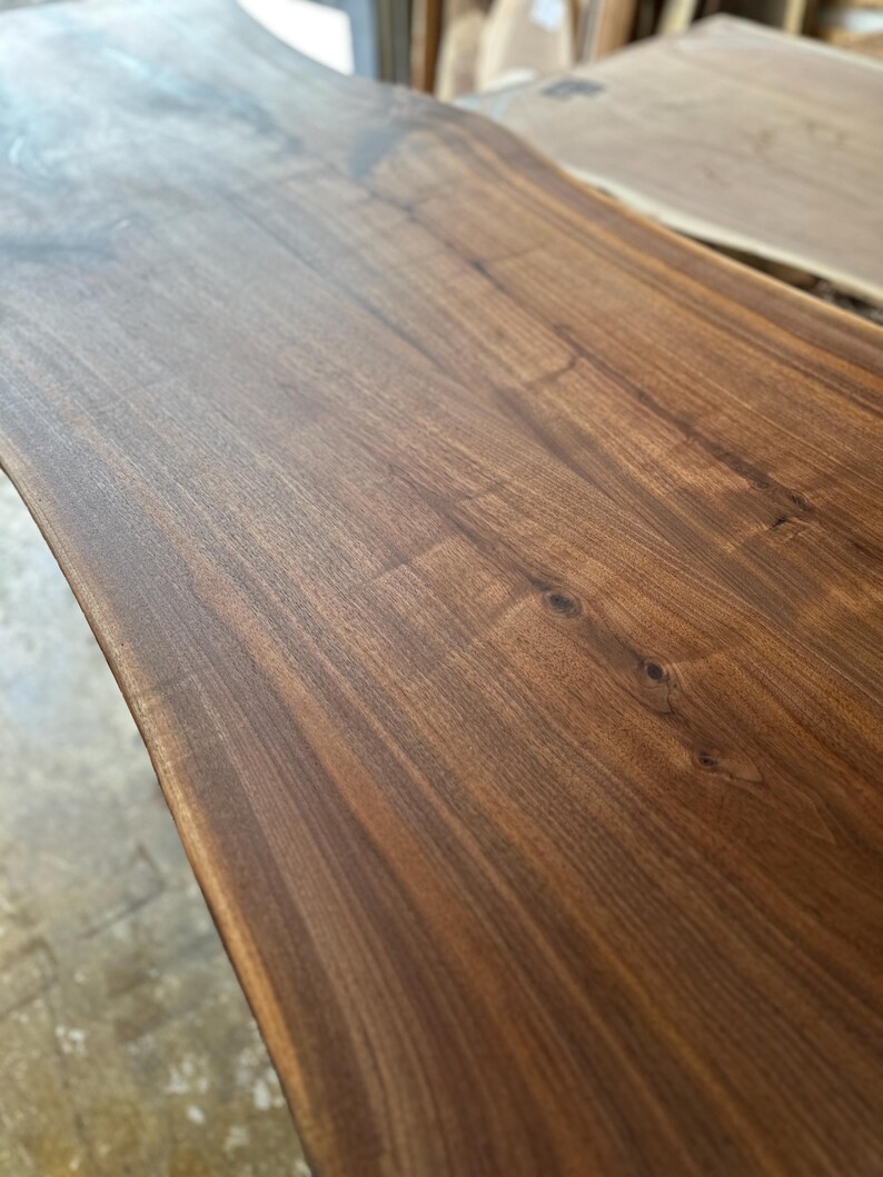May include: A rectangular wooden slab with a natural live edge, featuring a rich dark brown grain pattern with visible wood knots and a smooth, polished finish. The surface displays deep, warm tones and intricate organic wood textures.