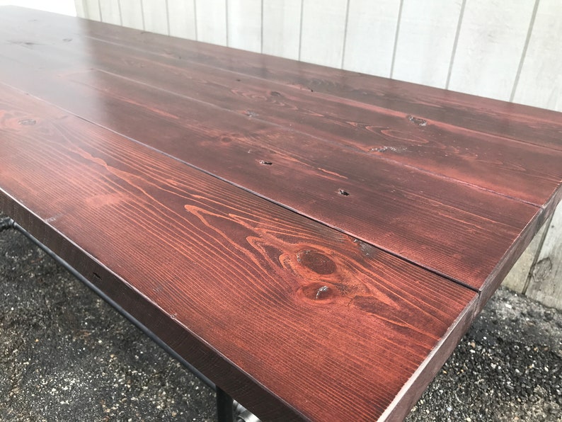 May include: A large, rectangular wooden table with a rich, dark reddish-brown stain. The tabletop is constructed from several planks, showcasing the natural wood grain. The table has metal legs.