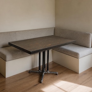 May include: A corner dining set featuring a dark wood table with a black metal base. The table is surrounded by a built-in bench with light gray cushions and a cream-colored base. The set is placed on a hardwood floor.