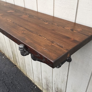 May include: A dark brown wooden shelf with a rustic finish is mounted to a white wall using black metal pipe brackets. The shelf is approximately 30 inches long.