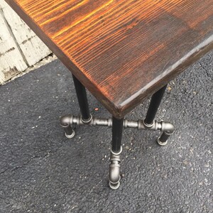 May include: A rustic wooden table top with a natural wood finish sits on a black metal pipe base. The table top has a live edge and is made of reclaimed wood.