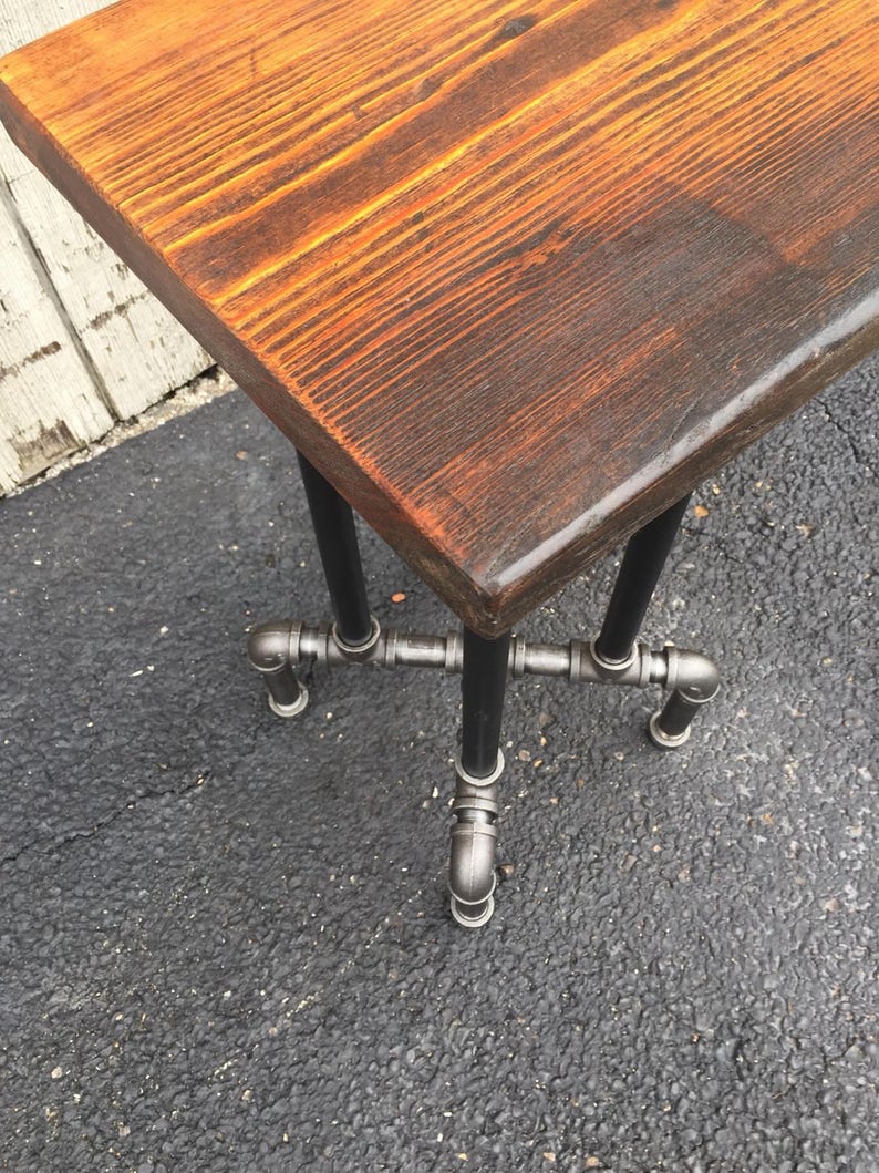 May include: A rustic wooden table top with a natural wood finish sits on a black metal pipe base. The table top has a live edge and is made of reclaimed wood.