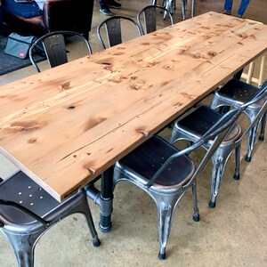 May include: A long, wooden table with a natural finish sits on a concrete floor.  The table is surrounded by six metal chairs with black seats.  The chairs have a distressed, vintage look.