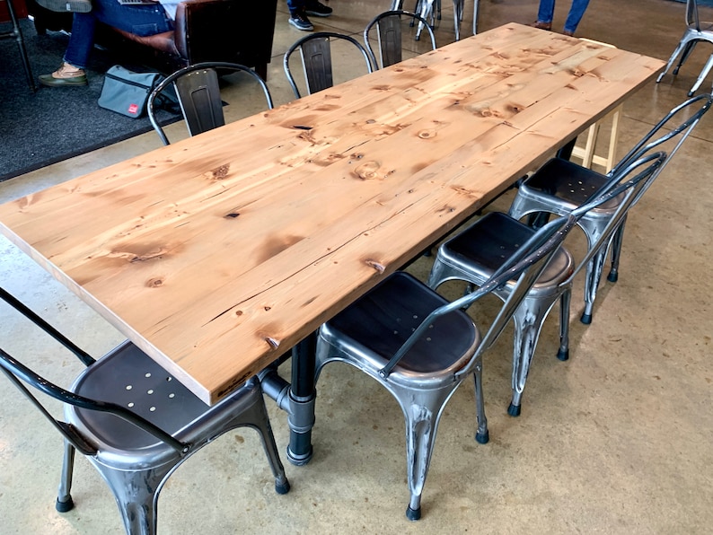 May include: A long, wooden table with a natural finish sits on a concrete floor.  The table is surrounded by six metal chairs with black seats.  The chairs have a distressed, vintage look.