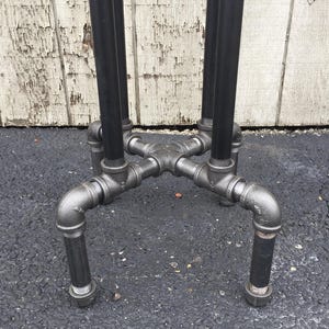 May include: Close-up of the base of a black metal table with four legs. The legs are made of pipe fittings and are connected to a central cross-shaped piece.