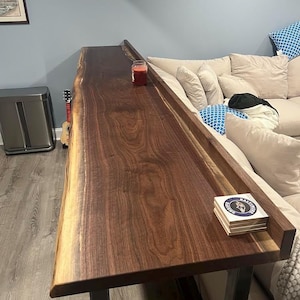 May include: A live edge wooden sofa table with a dark brown finish. The table has a natural edge and sits on black metal legs. A small candle and coasters are on the table.