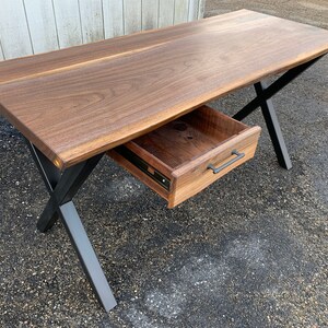 May include: A wooden desk with a live edge top and a single drawer. The desk has black metal legs in an X shape.